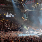 A performer executes a motorcycle stunt on a suspended track during an arena concert, captivating a packed audience.