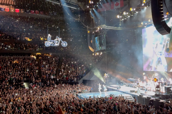 A performer executes a motorcycle stunt on a suspended track during an arena concert, captivating a packed audience.
