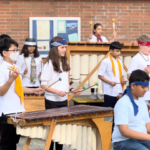 A diverse group of young musicians wearing headbands and scarves play marimbas and xylophones outdoors in front of a brick building.