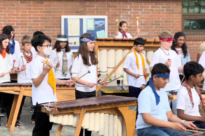 A diverse group of young musicians wearing headbands and scarves play marimbas and xylophones outdoors in front of a brick building.