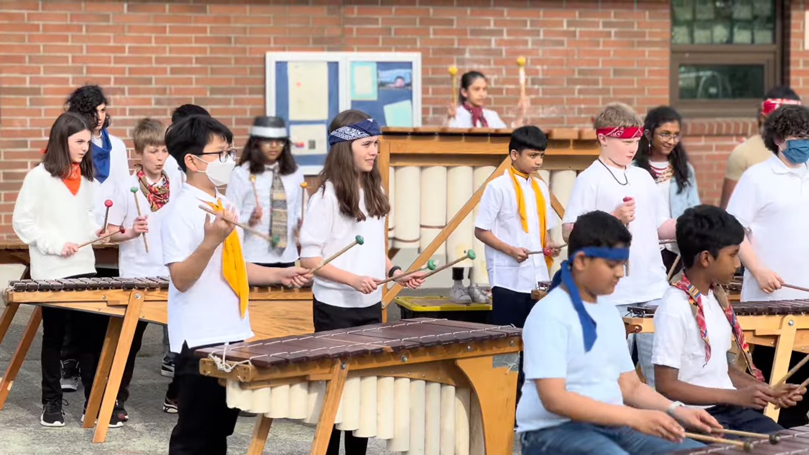 A diverse group of young musicians wearing headbands and scarves play marimbas and xylophones outdoors in front of a brick building.