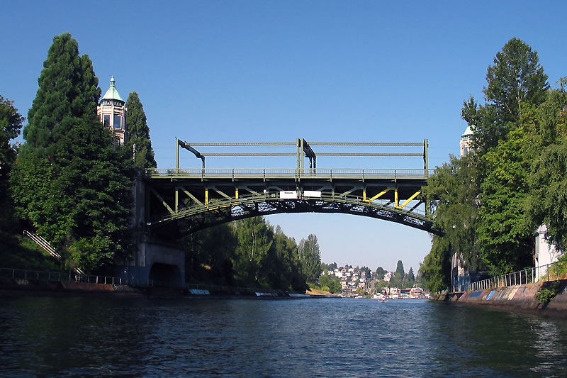 The Smith Street Bridge spans the Seattle River with lush green trees in the foreground, casting long shadows under the twilight sky.