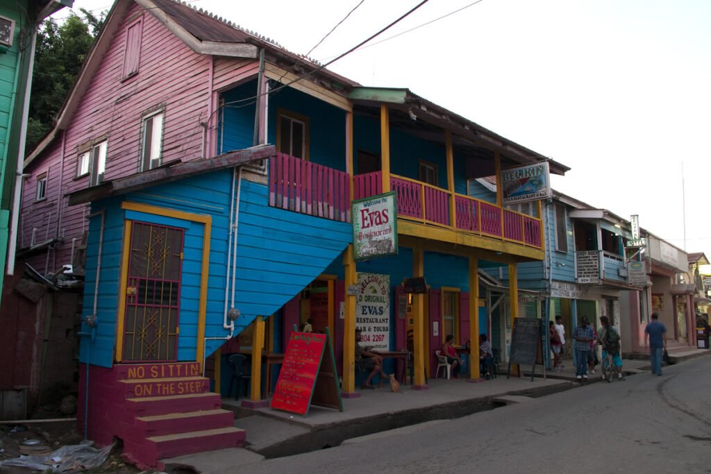 A street lined with colorful buildings featuring blue, pink, and yellow accents. Pedestrians walk along the sidewalk in front of small shops and restaurants.