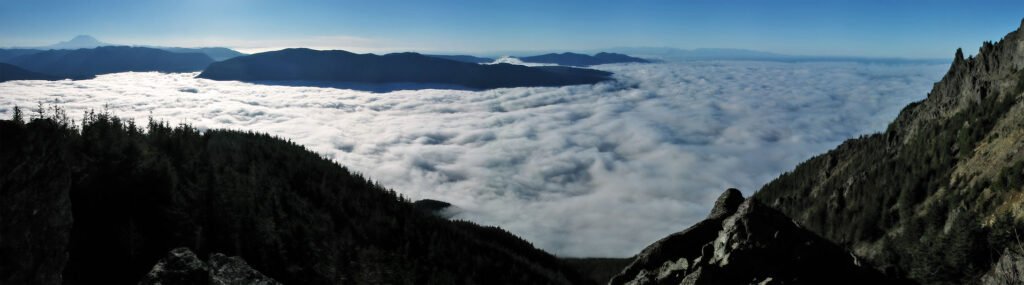 A breathtaking view from atop a mountain, looking down on a sea of clouds with distant mountains in the background. The sky is clear and blue, offering a serene and expansive vista.