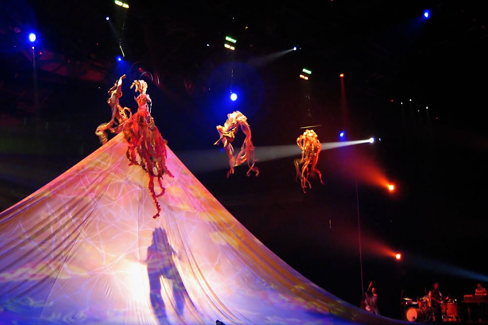 Acrobats performing on aerial silk against colorful lighting.