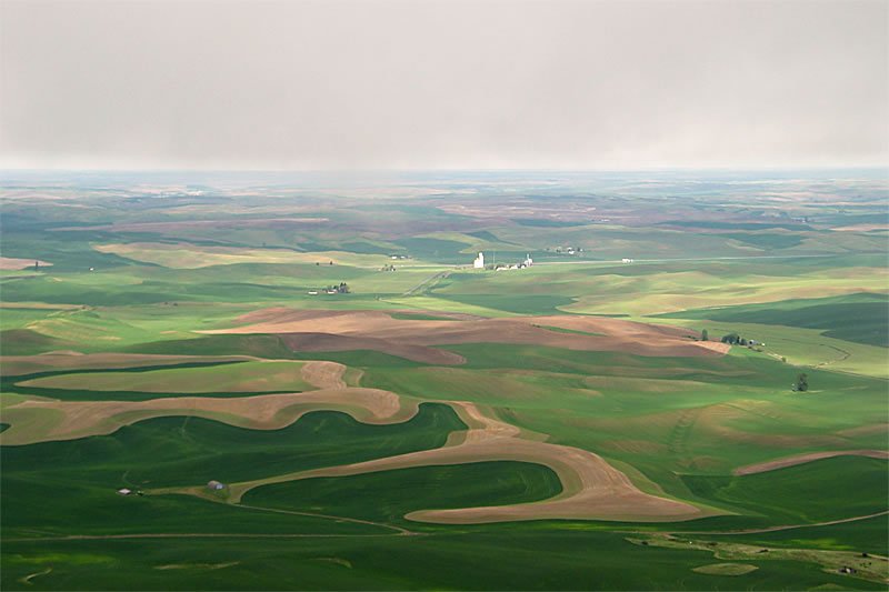 Lush green fields and winding dirt paths stretch across the hilly landscape under an overcast sky.