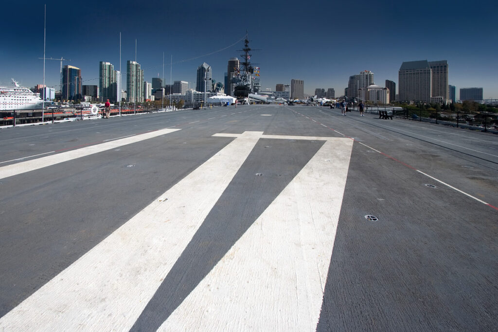 View of an aircraft carrier flight deck with white markings. In the distance, a city skyline is visible under a clear blue sky.