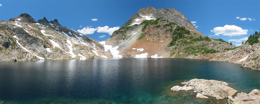A stunning panoramic view of Mt. Rainier with its alpenglow casting a warm hue over the granite granite and tundra landscape. A serene alpine lake reflects the beauty of the mountain.