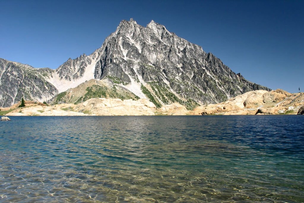 A tranquil mountain lake reflects the clear blue sky, bordered by rugged peaks and rocky shores.