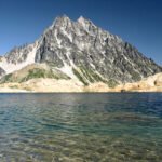 A tranquil mountain lake reflects the clear blue sky, bordered by rugged peaks and rocky shores.