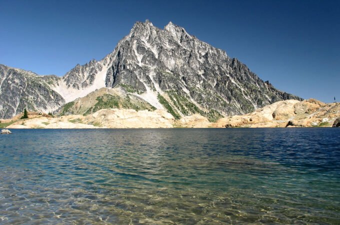 A tranquil mountain lake reflects the clear blue sky, bordered by rugged peaks and rocky shores.