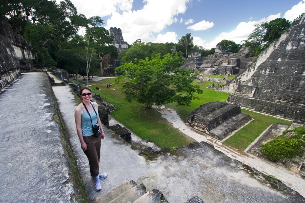 A woman stands on a stone pathway surrounded by ancient ruins and lush greenery under a bright blue sky.