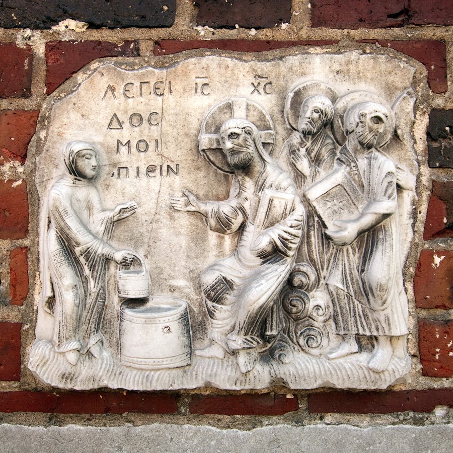 An intricate stone carving featuring multiple figures in traditional attire, engaged in various activities. The scene includes individuals pouring water and interacting with each other, set against a brick wall background.