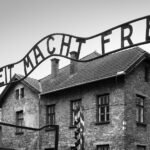 A black and white photograph of the infamous 'Arbeit Macht Frei' sign at the entrance of Auschwitz concentration camp.