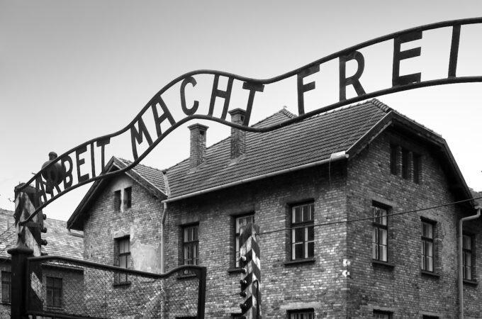 A black and white photograph of the infamous 'Arbeit Macht Frei' sign at the entrance of Auschwitz concentration camp.