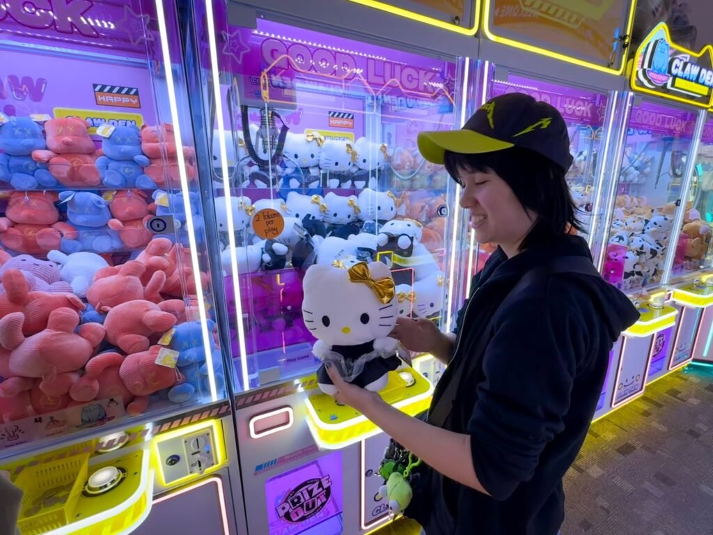 A girl in a black hoodie and cap is holding a Hello Kitty plush toy she won from a colorful claw machine at an arcade. The background shows various plush toys inside the arcade machines.