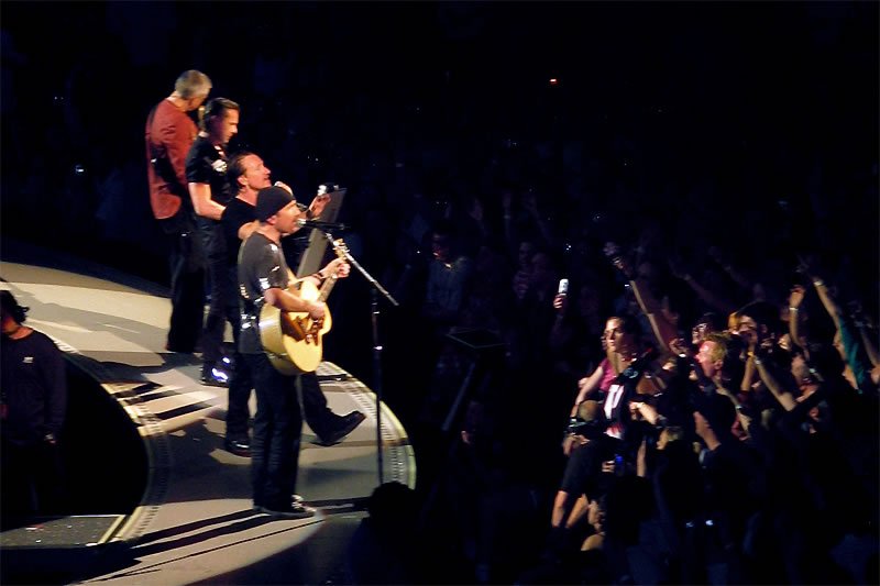 A band performs on stage with a crowd of people watching closely. Musicians play guitars and sing into microphones.