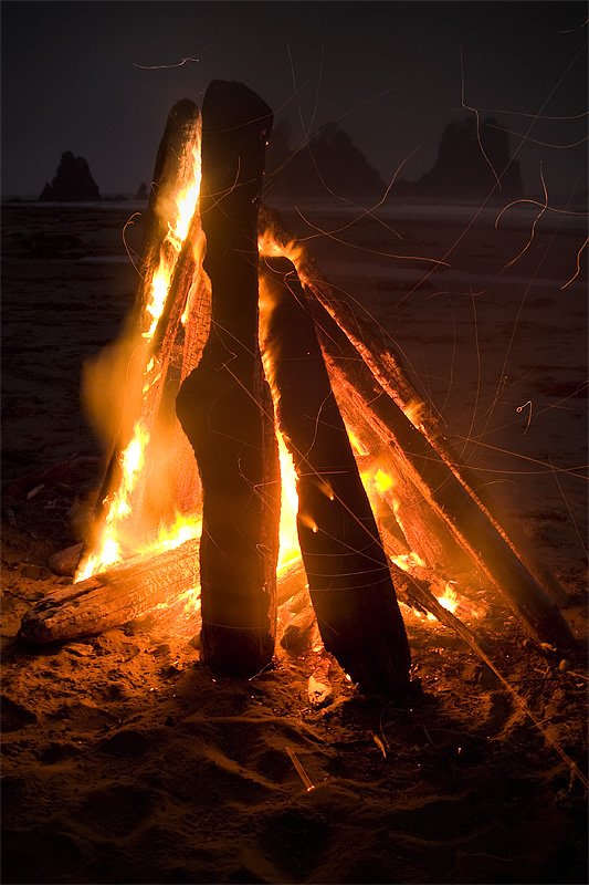 A cozy beach bonfire burns brightly against a darkening sky, casting warm light on the surrounding sand. The flames dance around wooden logs, creating a serene and inviting atmosphere.