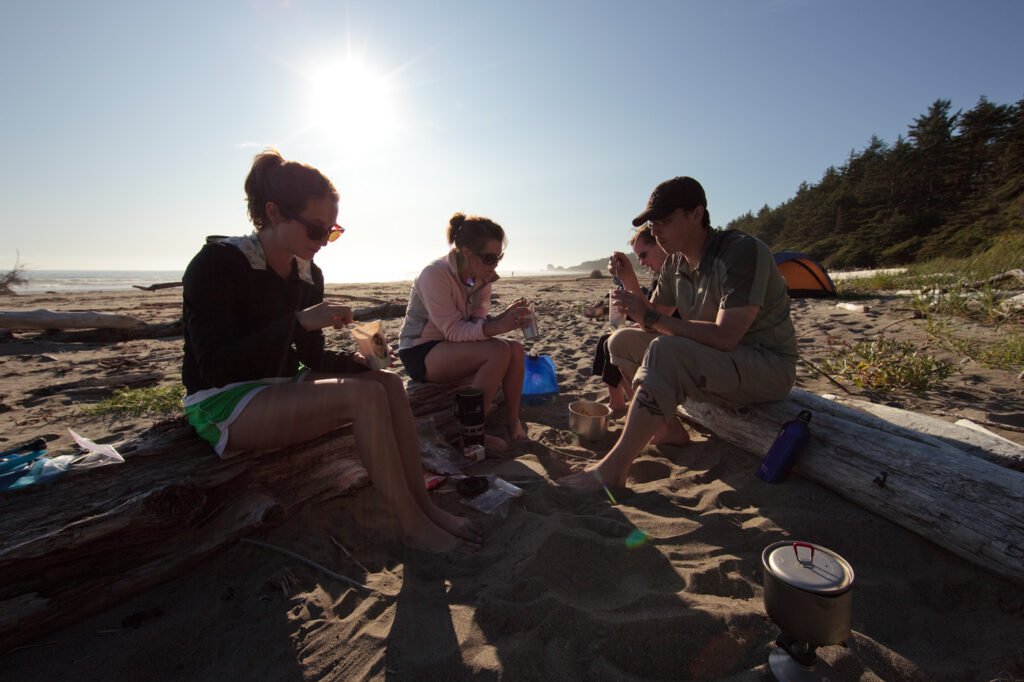 A group of friends enjoying breakfast on a sunny beach. The scene captures them sitting around a cooking pot on driftwood logs.