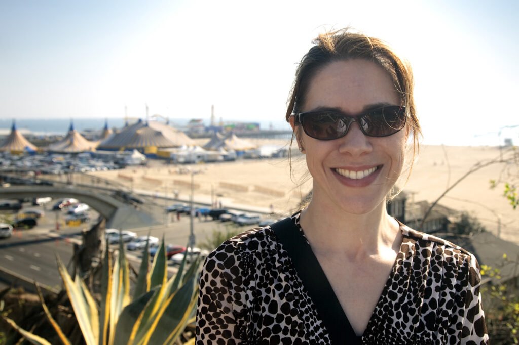 A woman wearing sunglasses smiles brightly with a beach and ocean in the background.