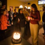 Dallas A group of friends gathered around a chocolate cake with lit candles in a cozy living room. The warm ambiance highlights their celebration.