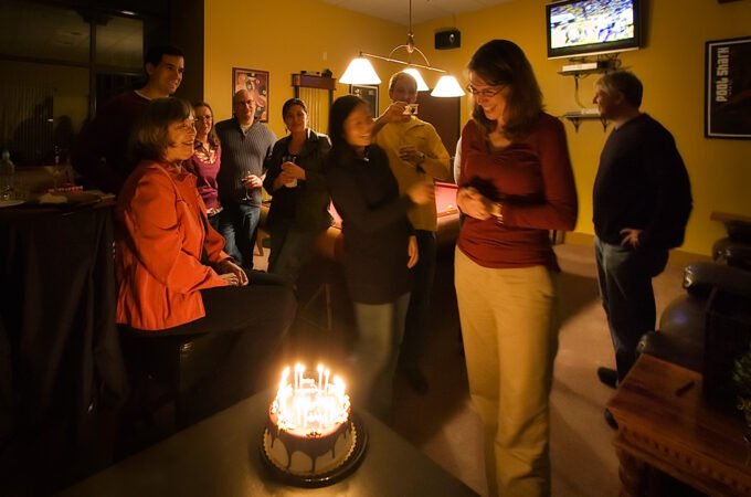 A group of friends gathered around a chocolate cake with lit candles in a cozy living room. The warm ambiance highlights their celebration.