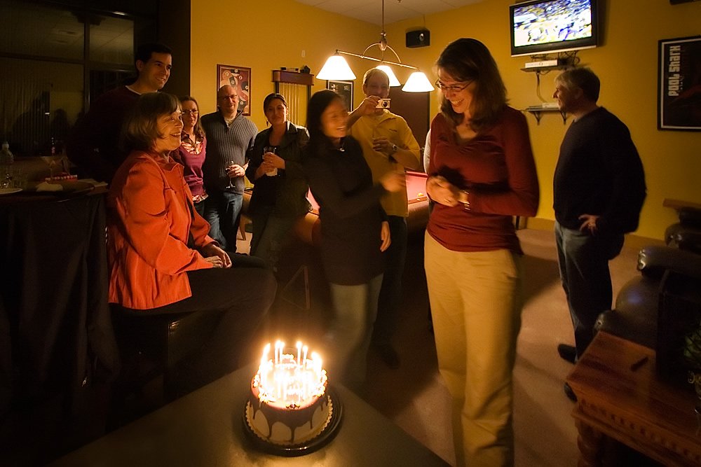 A group of friends gathered around a chocolate cake with lit candles in a cozy living room. The warm ambiance highlights their celebration.