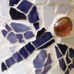 Close-up of a handmade vase featuring a dragonfly mosaic. The design uses broken blue ceramic tiles and a smooth amber glass stone for the eye.