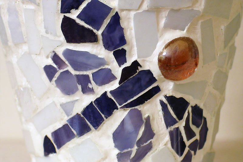 Close-up of a handmade vase featuring a dragonfly mosaic. The design uses broken blue ceramic tiles and a smooth amber glass stone for the eye.