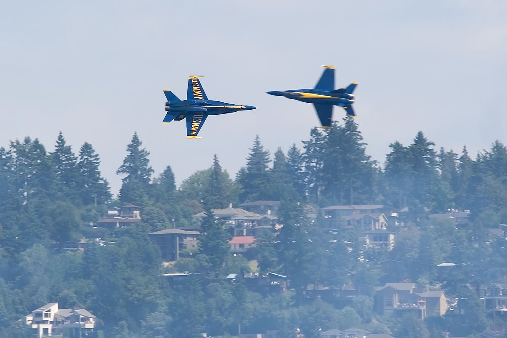 Two Blue Angels flying in formation over a residential area. The background features trees and houses typical of the Pacific Northwest region.