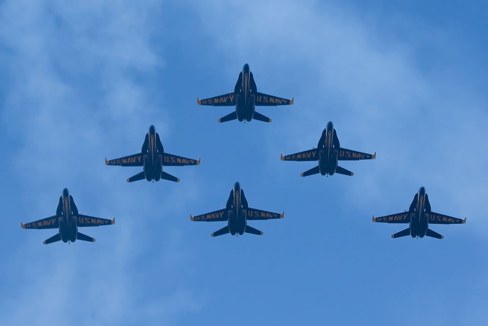 A formation of Blue Angels jets flying in a clear blue sky. The planes are positioned in a precise V-shape.