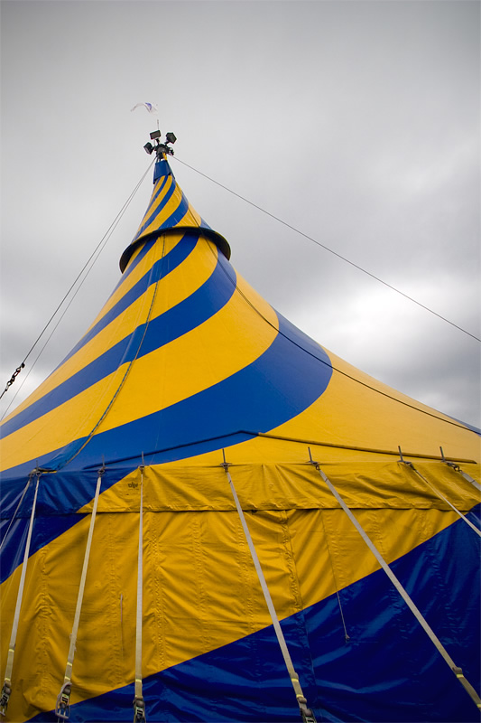 A vibrant blue and yellow striped circus tent stands tall against an overcast sky. The tent's fabric is stretched taut, showcasing its striking colors.