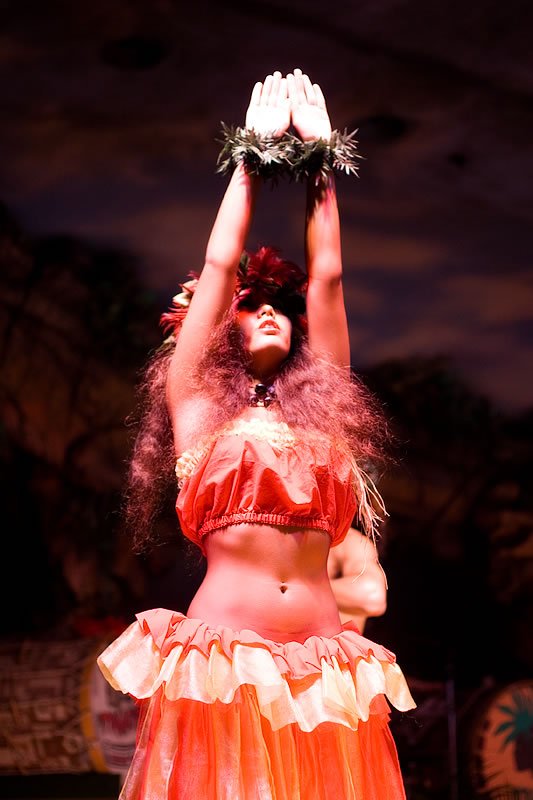 Dancer performing with a blush-red costume, standing stage-left with arms raised rightward. Looks like they're holding a decorative prop above their head.