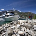 Campbell’s Resort A boat is docked at a rocky shoreline with snow-capped mountains in the background under a clear blue sky.