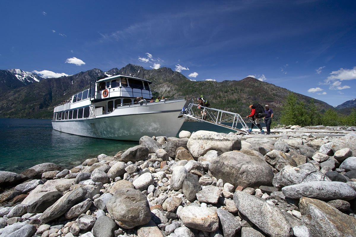 A boat is docked at a rocky shoreline with snow-capped mountains in the background under a clear blue sky.