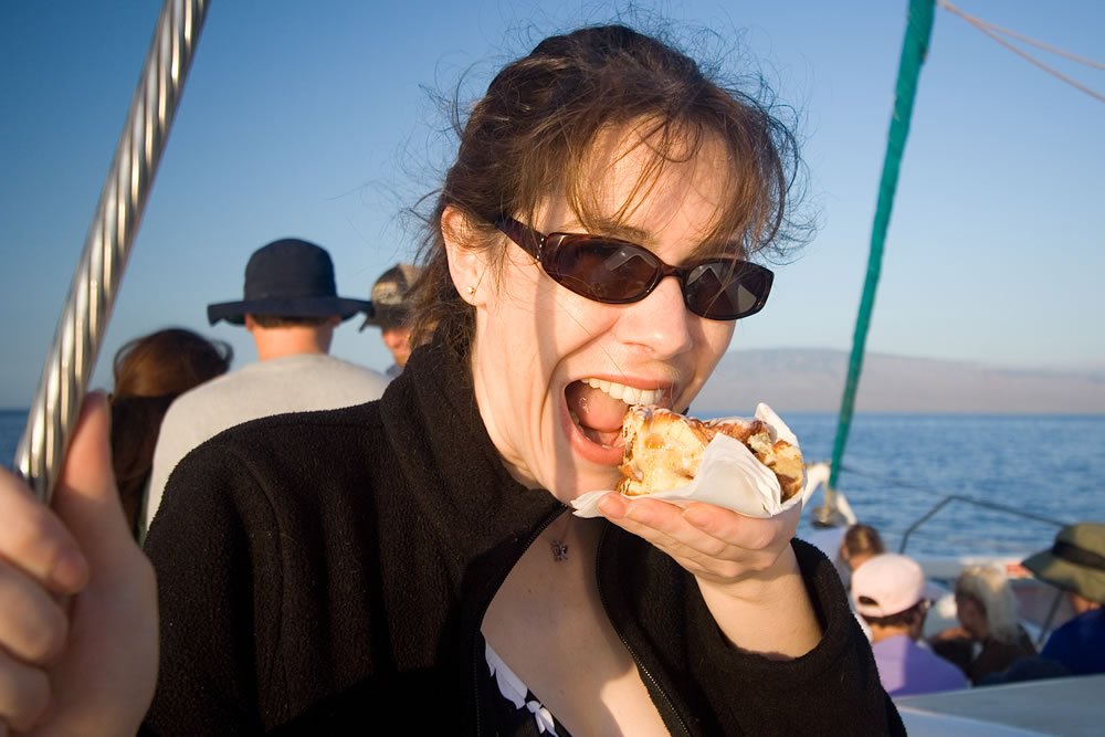 A woman wearing sunglasses enjoys a snack on a boat with others in the background. The scene showcases a sunny day on the water.