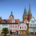 Historic half-timbered houses lined up along a riverfront with twin spires towering behind under a clear blue sky.