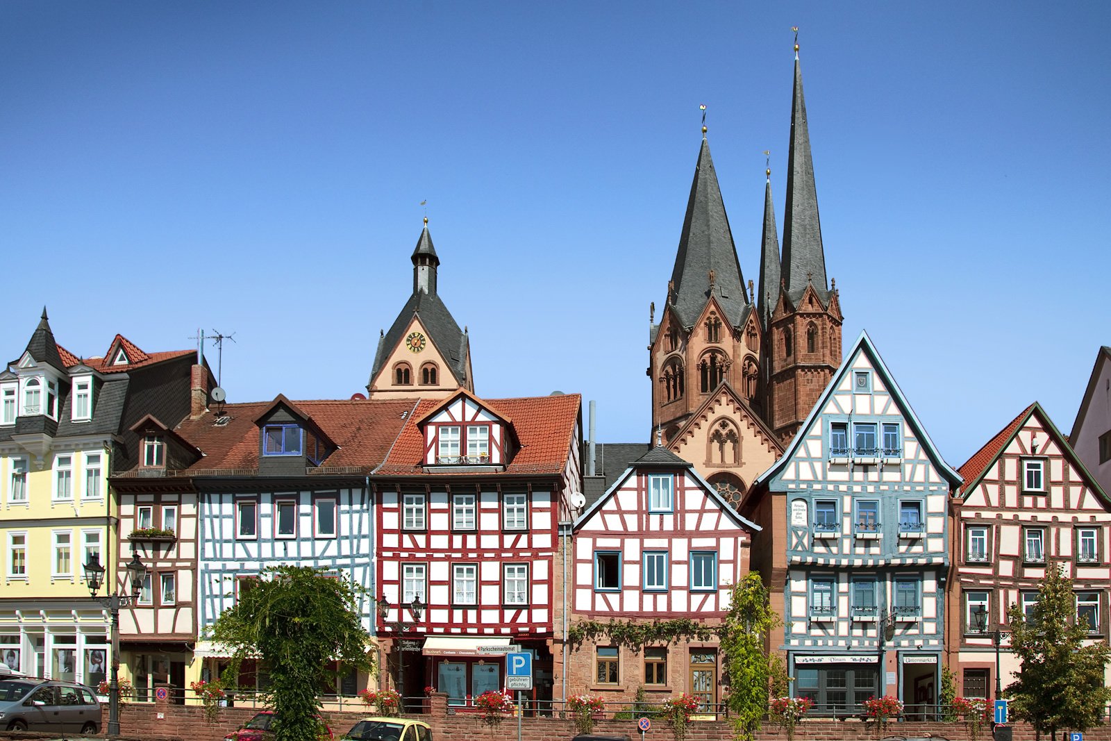 Historic half-timbered houses lined up along a riverfront with twin spires towering behind under a clear blue sky.