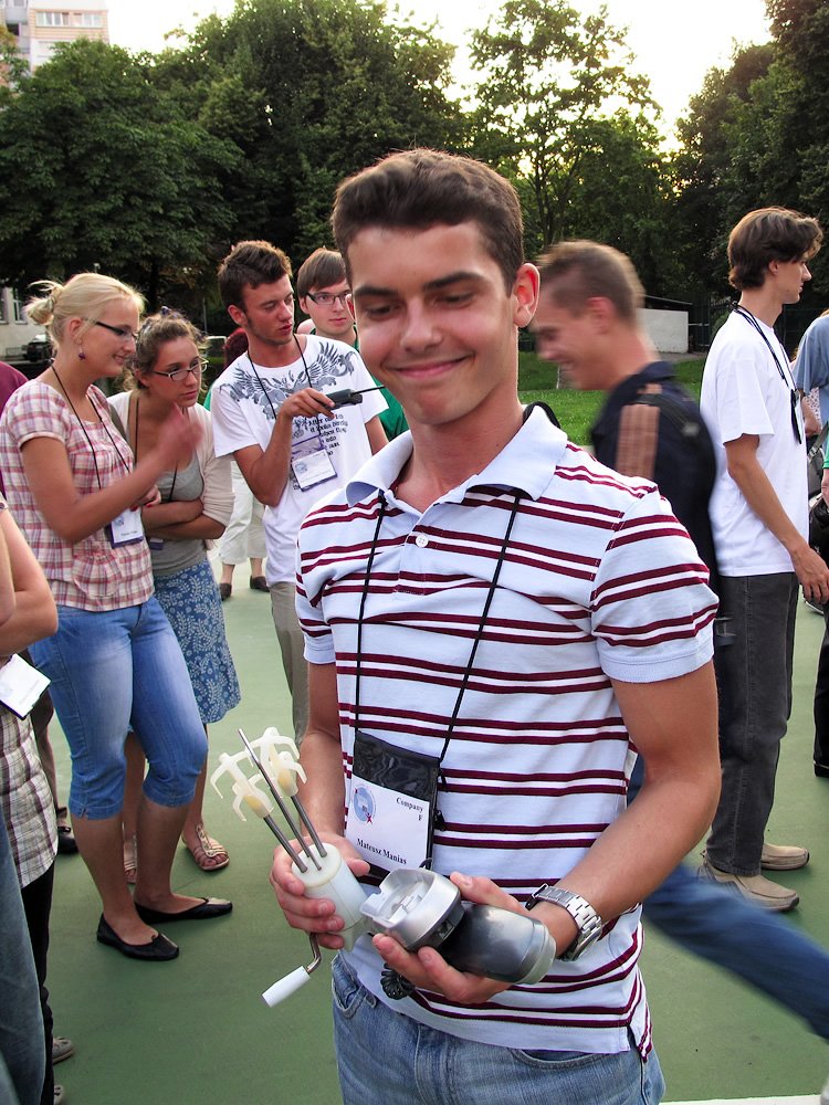 A young man holds bowling pins and a ball at an outdoor event. He is surrounded by friends enjoying a casual gathering.
