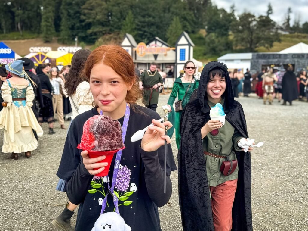 Two children at an outdoor medieval fair; one holds a red slushie and the other a blue ice cream.