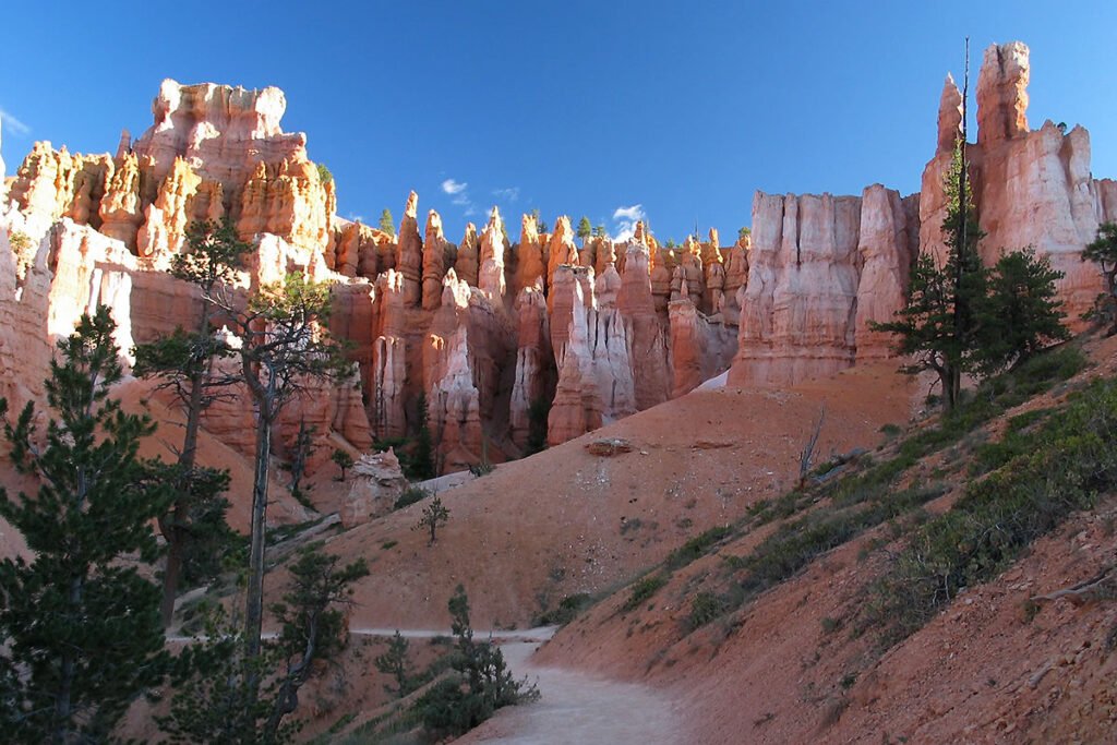 A scenic view of Bryce Canyon with reddish-orange rock formations and a clear blue sky. A winding trail is visible, flanked by green vegetation.