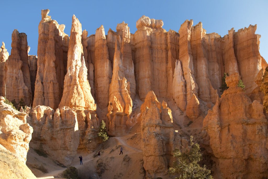 Towering, orange rock formations rise majestically against a clear blue sky. A lone figure walks along the trail, dwarfed by the grandeur of nature.