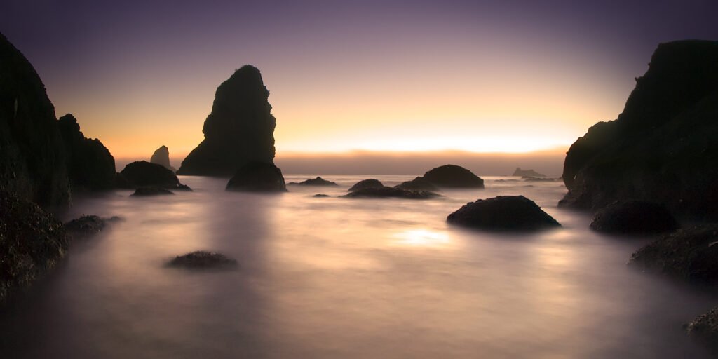 A serene sunset scene at Cannon Beach with Haystack Rock silhouetted against a vibrant sky. The calm waters reflect the warm hues of the setting sun.