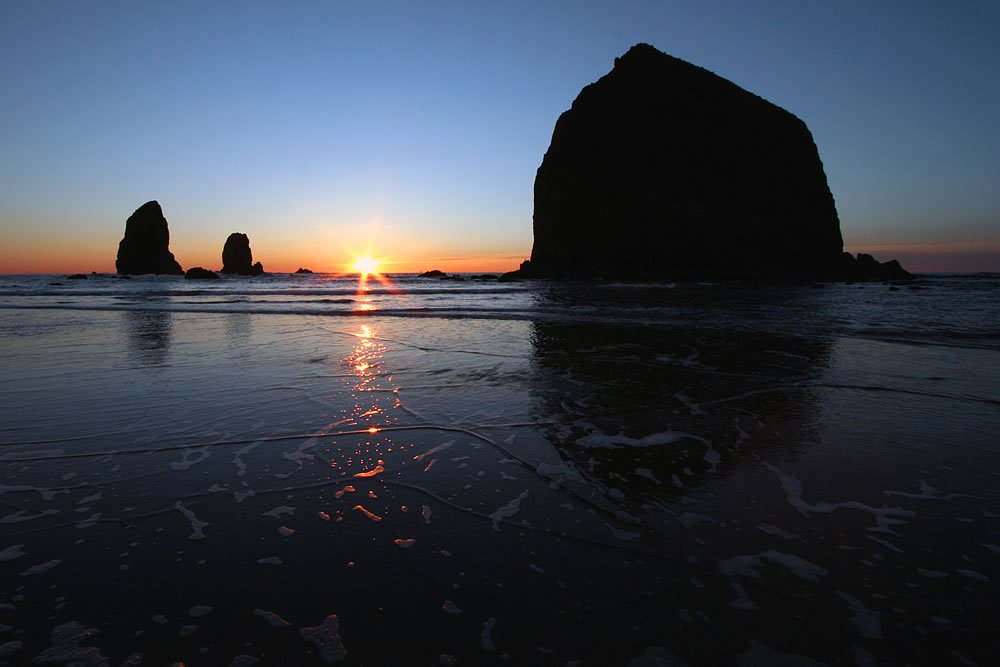 Silhouetted rock formations against a vibrant sunset sky with reflections on the wet beach.