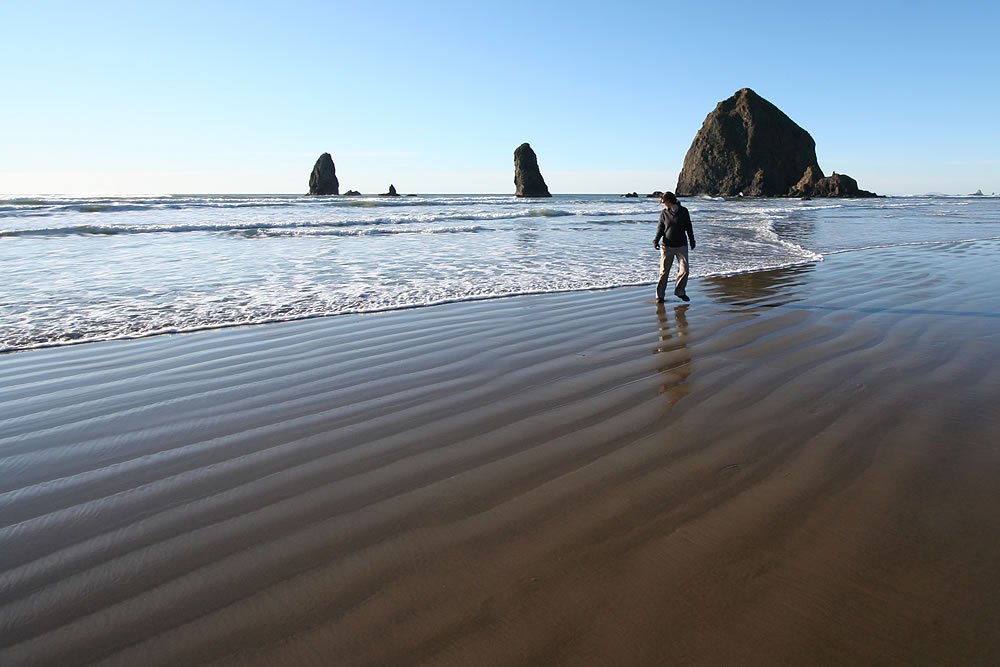 A person strolls along the wet sands of Cannon Beach with Haystack Rock in the background.