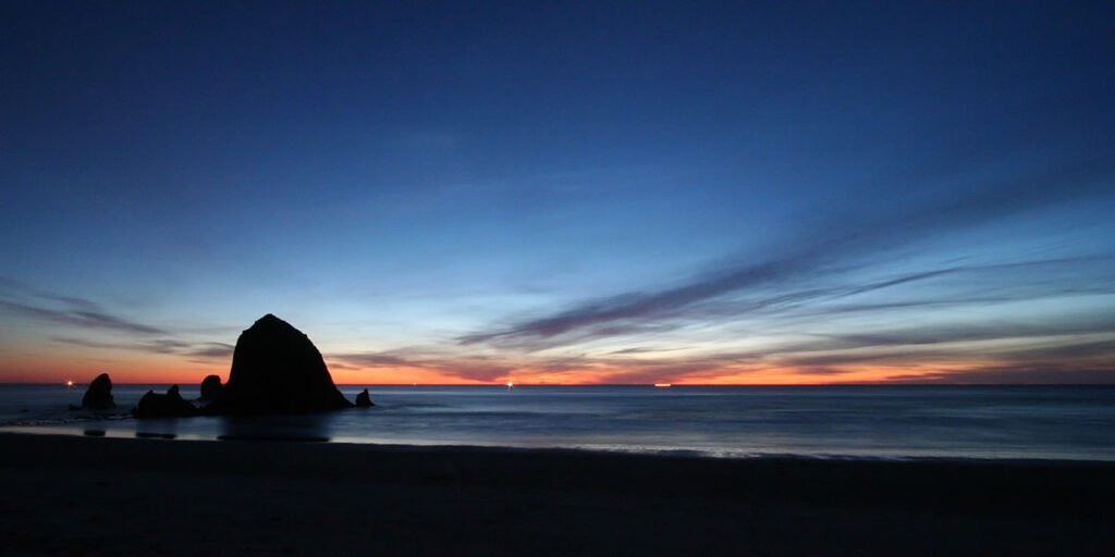 Serene sunset view of the Pacific Ocean with Haystack Rock silhouetted against a vibrant blue and orange sky. Ideal scenery enjoyed by John and Kristie during their travels.