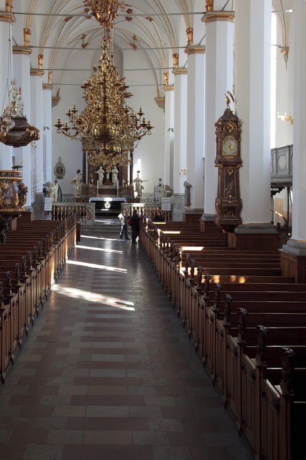 A serene view inside a grand cathedral featuring tall white pillars, ornate chandeliers, and wooden pews arranged neatly along a central aisle.