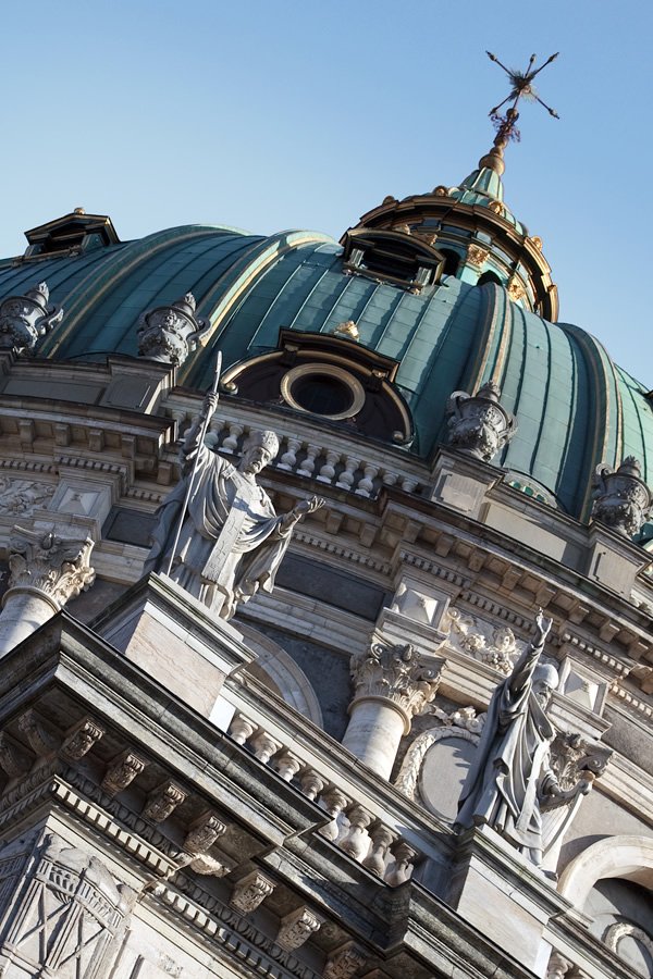 A close-up view of an ornate cathedral dome with statues and a cross against a clear blue sky.