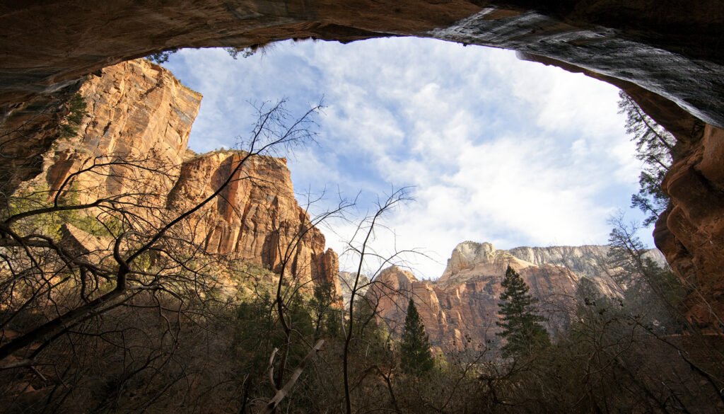 A stunning view of towering red rock cliffs framed by a natural cave opening. Sparse vegetation and blue sky add to the scene.
