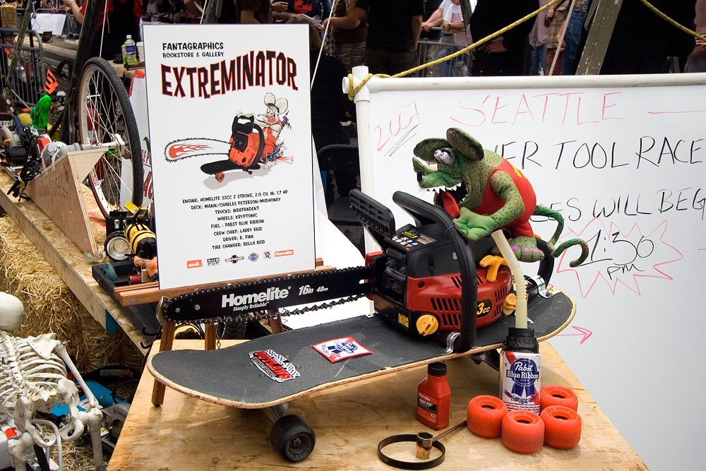 A skateboard equipped with a chainsaw is set up on a track, alongside signs advertising the Extreme Motor Tool Race.